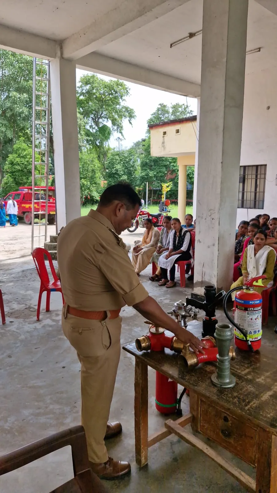 Fire officer demonstrating hydrant couplings and a dry powder extinguisher to seated trainees